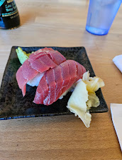 A black plate holds neatly sliced pink sashimi with shredded daikon, shiso leaves, and pale pickled ginger on a wooden table.