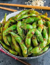 A bowl of bright green edamame pods sprinkled with red chili flakes, with wooden chopsticks resting behind it on a dark tabletop.