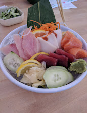 A colorful white bowl holds assorted sashimi, shrimp, shredded carrots, cucumber, broccoli, pickled ginger, and green garnish on a wooden table.