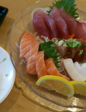 A glass platter of colorful sashimi, including salmon and tuna, with shredded daikon, green herbs, lemon wedges, and a fresh, appetizing presentation.