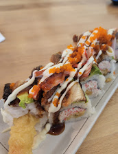 A long sushi roll on a rectangular plate, drizzled with creamy white sauce and dark glaze, topped with orange roe and crunchy tempura crumbs.
