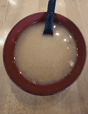 A red-rimmed bowl holds cloudy beige liquid on a wooden surface, with a dark-handled spoon standing inside; soft reflections add a calm tone.