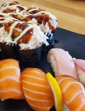 Assorted sushi on a black slate salmon nigiri in front, pale fish slices beside it, and saucy, mayo-drizzled rolls behind on a wooden table.