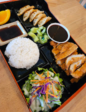 Bento box on a wooden table with white rice, dumplings, sushi rolls, fried chicken, salad, dipping sauces, and an orange slice.