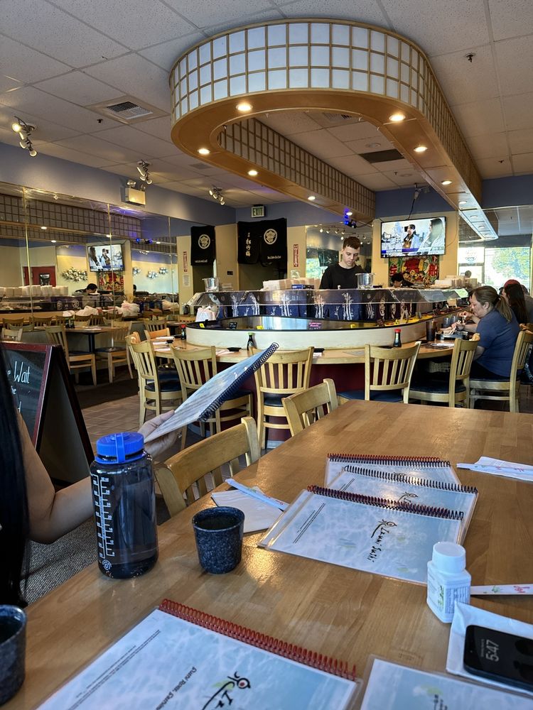 Bright Japanese restaurant interior with a curved illuminated ceiling, central sushi bar, diners, wooden tables, menus and water bottles in foreground.