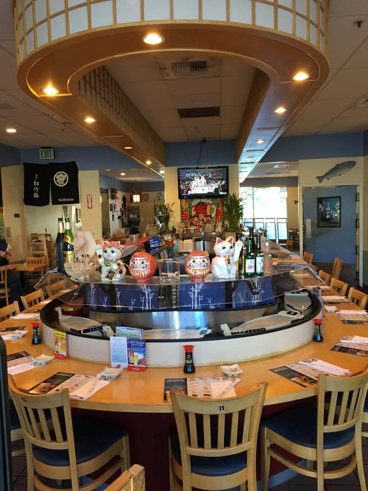 Bright, welcoming Japanese restaurant with a curved sushi bar, colorful lucky cats, bottled drinks, menus, and wooden chairs beneath warm lights.