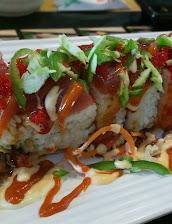 Close-up of a sliced sushi roll on a white plate, topped with cilantro, avocado, tomato, and drizzled with spicy red and creamy sauces.