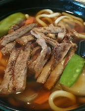 Close-up of noodle soup topped with sliced grilled beef, green beans, carrots, and onions in rich broth, served in a dark bowl.