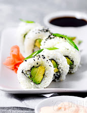 Close-up of sliced sushi rolls with avocado and cucumber, topped with sesame seeds and herbs, beside pickled ginger and soy sauce.