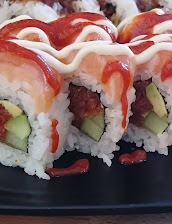 Close-up of sushi rolls topped with red fish and white sauce, filled with avocado and cucumber, arranged on a black plate against a dark background.