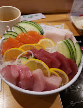 Colorful bowl of assorted sashimi—salmon, tuna, and white fish—with cucumber, lemon, and onion garnishes on a dining table; appetizing.