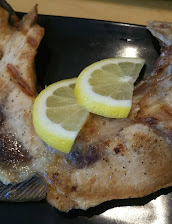 Cooked whole fish on a black plate, topped with two lemon slices; browned, flaky skin and fins create a rustic, appetizing dish.