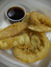 Golden-brown fried seafood with crisp, textured batter piled on a white tray beside a small cup of dark dipping sauce.