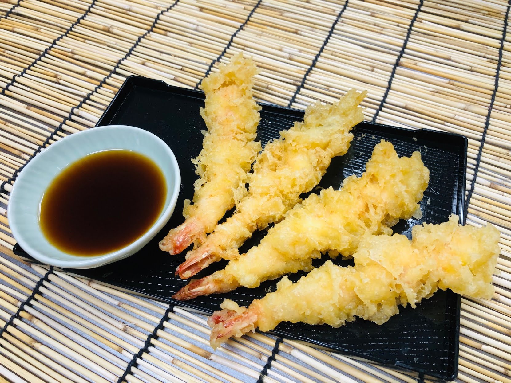 Golden shrimp tempura on a black plate with dark dipping sauce, set on a bamboo mat; a crisp, appetizing Japanese meal.