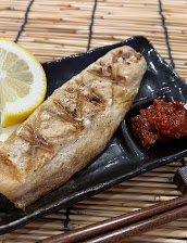 Grilled fish fillet with browned seasoning on a black plate, served with a lemon wedge and red sauce, against a bamboo mat with chopsticks.