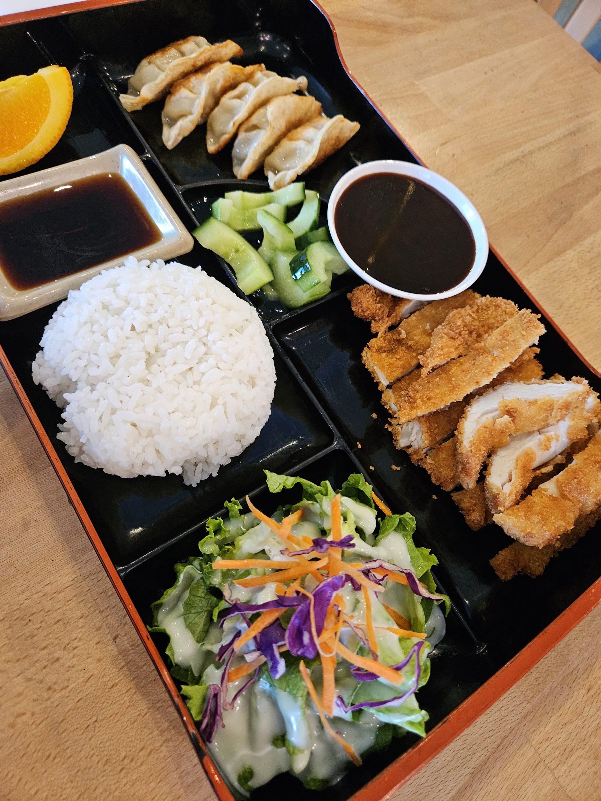 Japanese bento box with white rice, crispy fried chicken, dumplings, salad, cucumber, orange, and dipping sauces, appetizingly arranged.