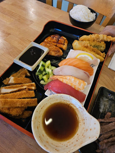 Japanese bento meal with sushi, tempura, fried cutlets, grilled fish, rice, pickles, and soy sauce on a wooden table; inviting feast.