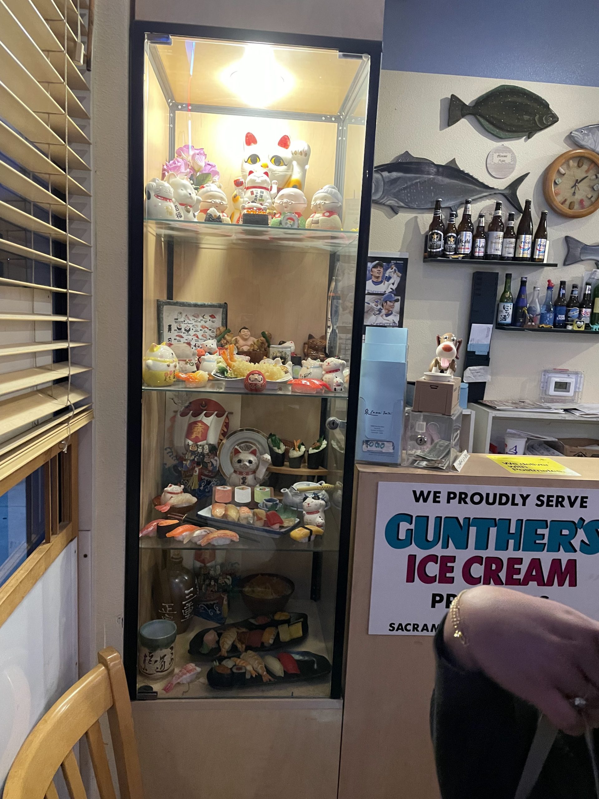 Restaurant counter with a glass cabinet of colorful Japanese ornaments, mounted fish, drinks, and a sign promoting Gunther’s ice cream.