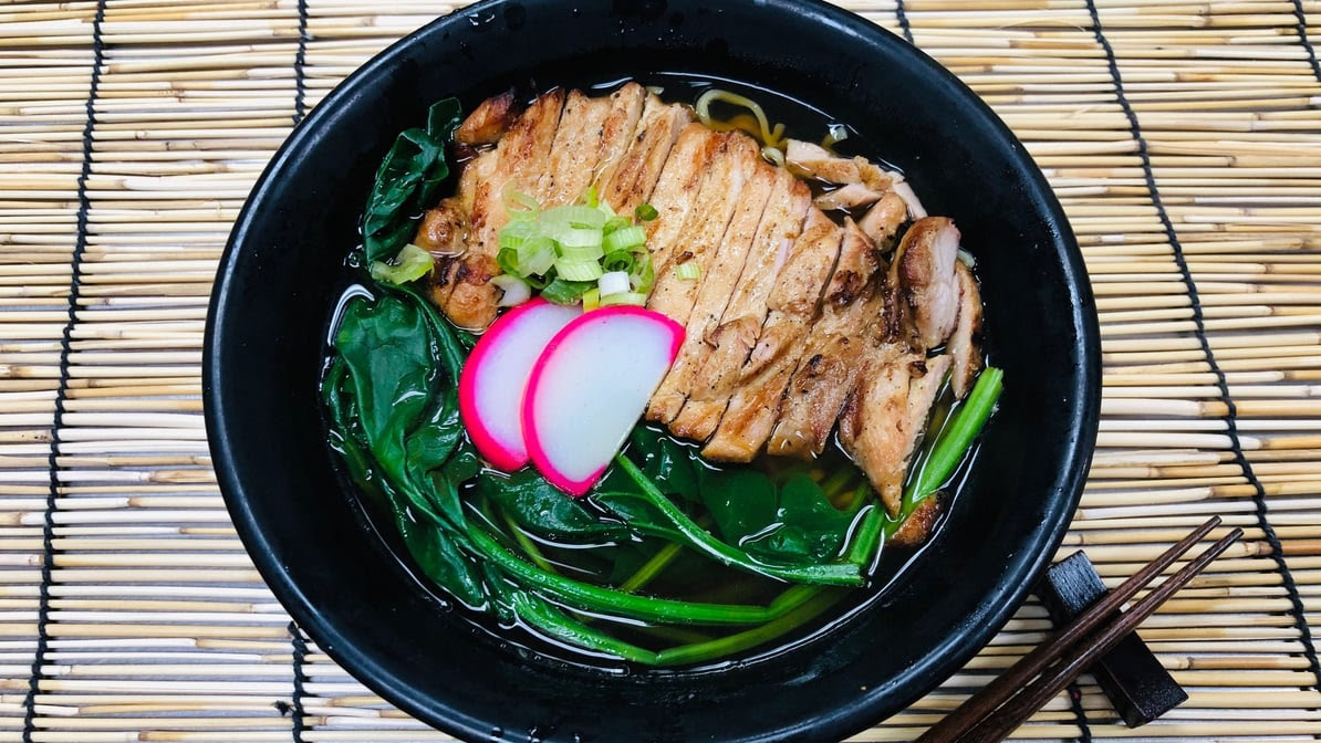 Top-down view of a black bowl of ramen with sliced grilled chicken, leafy greens, scallions, and radish in broth, with chopsticks beside it.