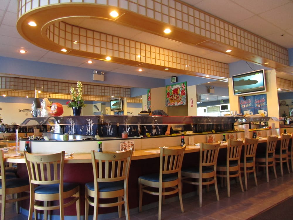 Warmly lit Japanese restaurant interior with sushi bar, wooden stools, decorative lanterns, plants, artwork, and overhead menu screens.