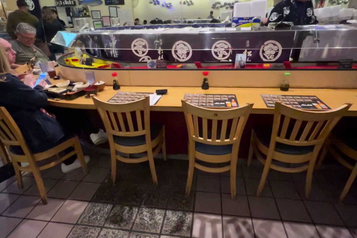 Busy sushi restaurant with chefs serving diners at a counter, aquarium tanks glowing blue behind them, and wooden chairs and menus in front.
